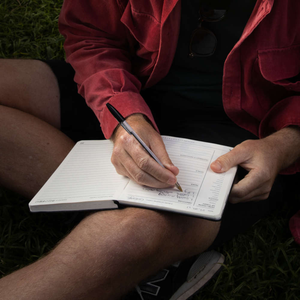 A person sitting outdoors on grass, writing in a journal for men with a black pen. The person is wearing a red jacket over a black shirt and dark shorts.
