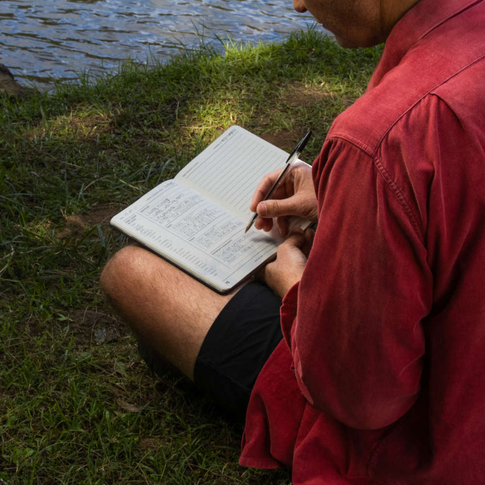 A man sitting outdoors by the water, writing in an open journal with a pen. The person is wearing a red long-sleeve shirt and black shorts, with grass and water visible in the background.