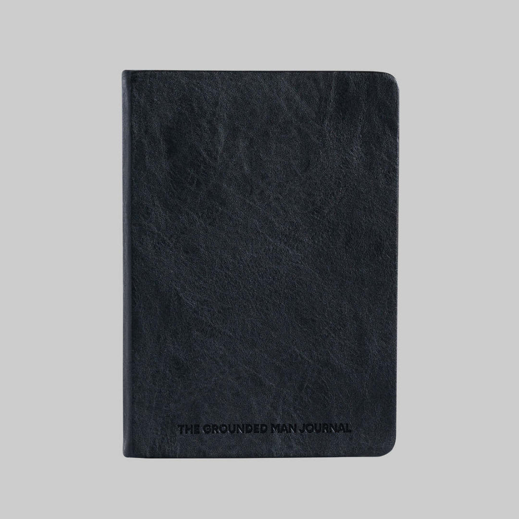 A black textured leather journal with 'THE GROUNDED MAN JOURNAL' embossed on the front, shown against a solid gray background.