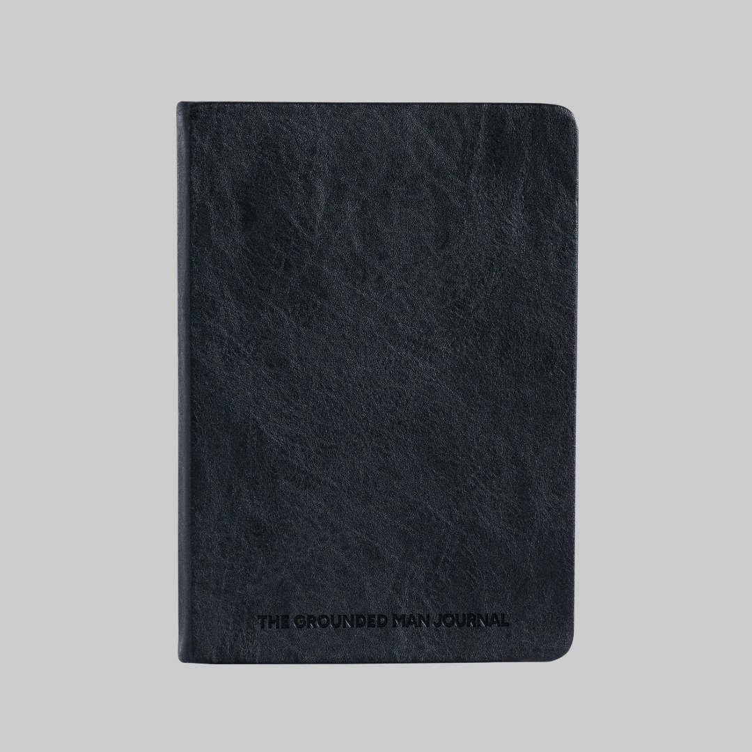A black textured leather journal with 'THE GROUNDED MAN JOURNAL' embossed on the front, shown against a solid gray background.