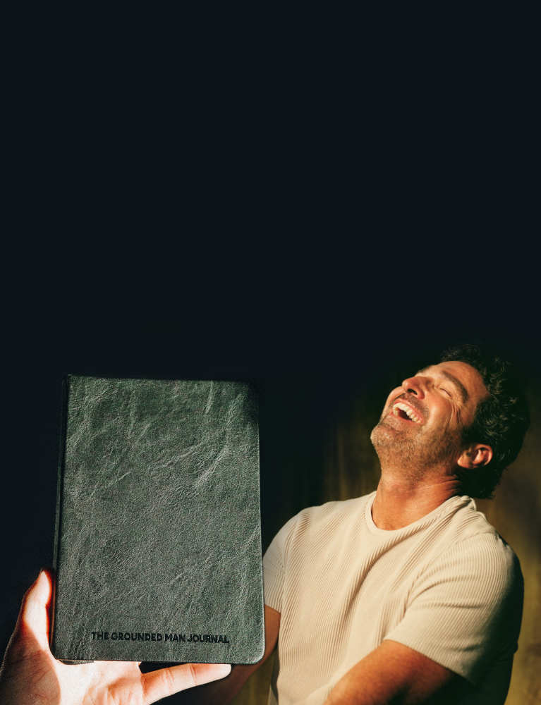 A man in a white t-shirt holds up a dark gray textured journal with the title 'THE GROUNDED MAN JOURNAL' visible on the cover against a dark background.