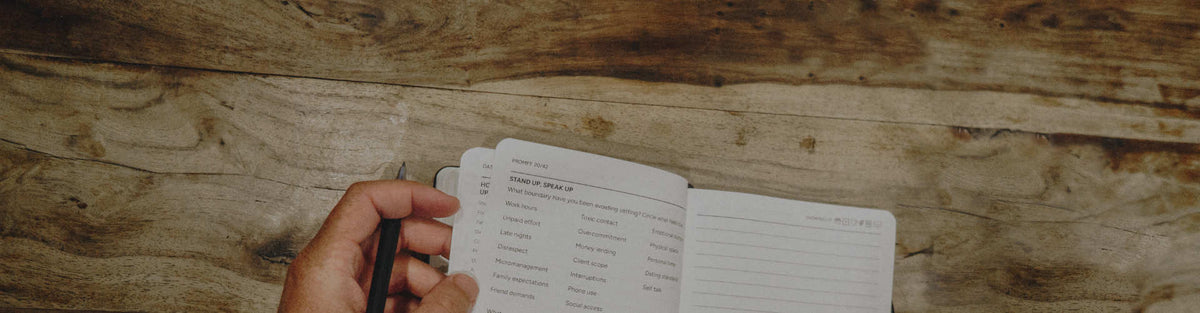 A hand holding a black pen over an open Grounded Man Journal on a rustic wooden tabletop.