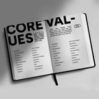 An open hardcover journal or notebook spread on a neutral gray background, featuring large black title text reading 'CORE VALUES' and columns of smaller words and phrases on both pages.