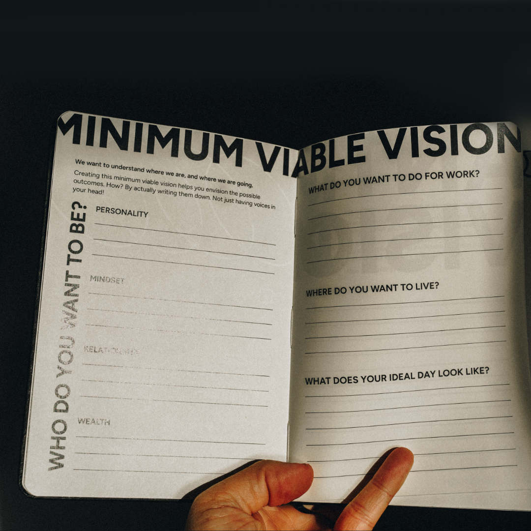 An open notebook or journal spread on a dark background, showing printed prompts such as 'MINIMUM Viable VISION' and 'WHO DO YOU WANT TO BE?' with lined writing sections. A hand is holding the lower edge of the pages.