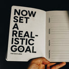 An open notebook or journal held by a hand, showing a left page with the bold text 'NOW SET A REALISTIC GOAL FOR EACH AREA' and a right page with lined sections labeled FAMILY, SOCIAL, HEALTH, and FINANCIAL.